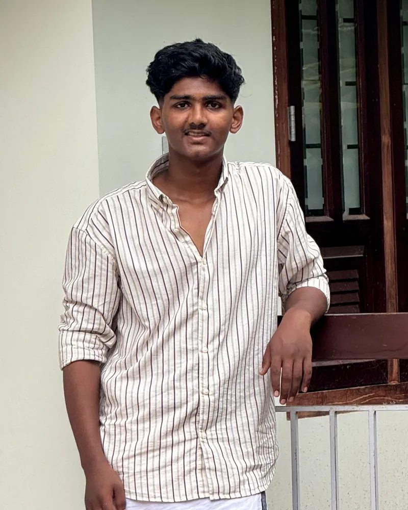 Portrait of Rishan KP leaning on a wooden railing, wearing a striped shirt and a white mundu